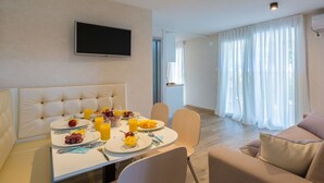 Un salon moderne comprenant un coin repas, un téléviseur à écran plat fixé au mur et une grande fenêtre ornée de rideaux légers.