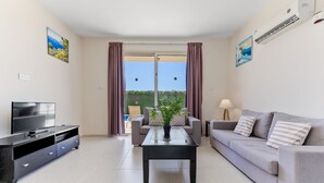 Ein modernes Wohnzimmer mit Flachbildfernseher, einer Couch, einem Couchtisch und Blick ins Freie durch große Fenster.