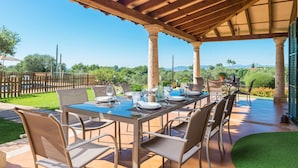Une terrasse extérieure avec une longue table dressée pour le repas, entourée de chaises, et protégée par une pergola en bois offrant une vue dégagée sur le paysage.