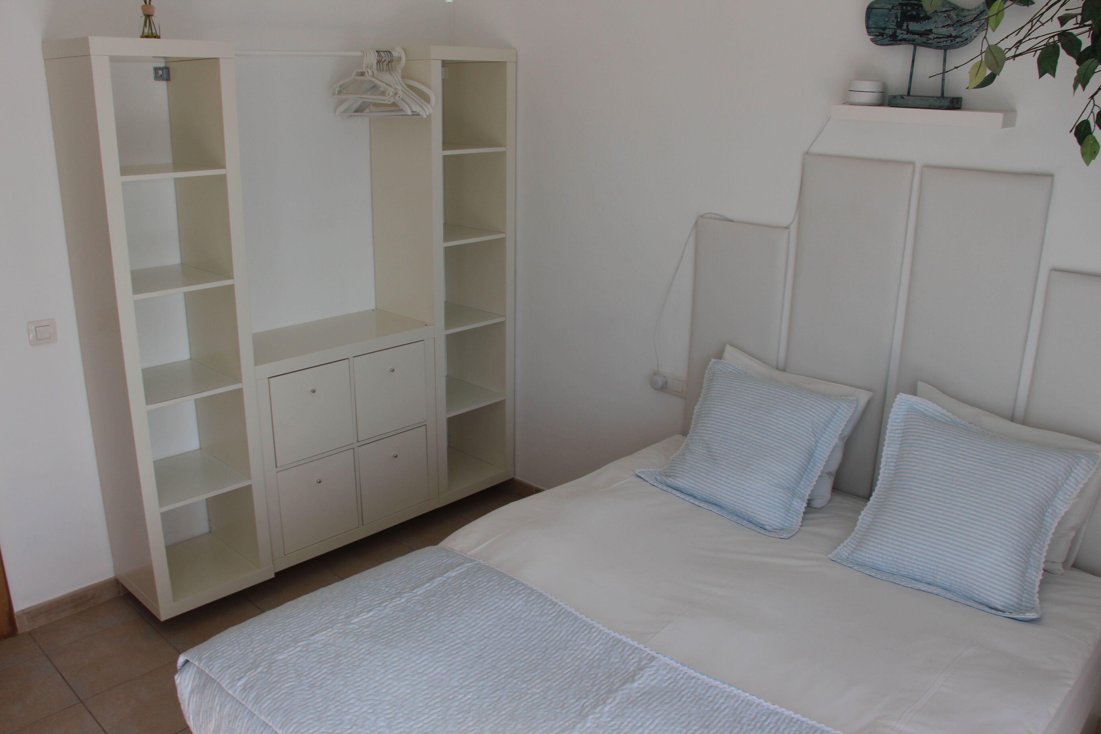 A bedroom with a bed, two pillows, a white wardrobe, and a small shelf with a plant.