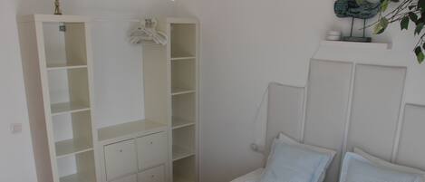 A bedroom with a bed, two pillows, a white wardrobe, and a small shelf with a plant.