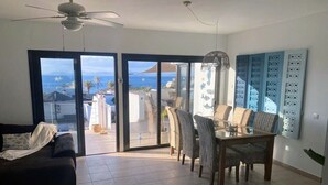 A living room with a sofa, dining table, chairs, and a ceiling fan. There is a sliding glass door leading to a balcony with a view of the ocean. - Luxury Villa with Private Heated pool, FREE WiFi,  (Playa Blanca, Yaiza)