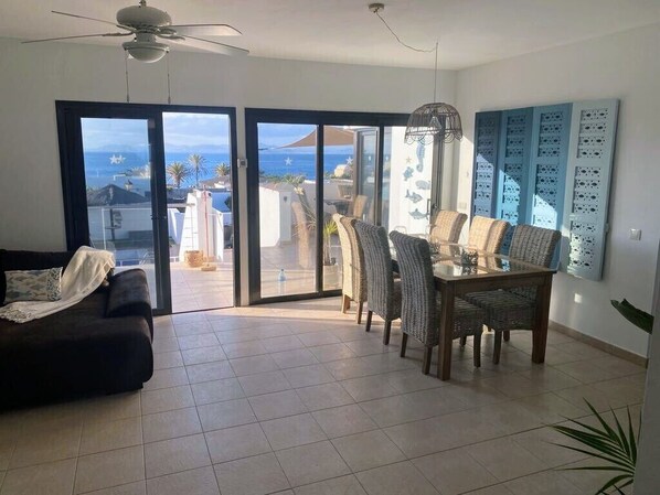 Sala de estar com sofá, mesa de jantar, cadeiras e ventilador de teto. Há uma porta de vidro deslizante que dá para uma varanda com vista para o oceano.