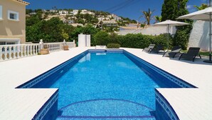Une piscine rectangulaire bordée de carreaux, entourée d’une terrasse blanche. On y trouve des chaises longues et des parasols, et la vue s’étend sur une colline avec des bâtiments en arrière-plan.