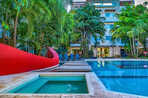 A pool area with lounge chairs and a red structure, surrounded by palm trees and a building with multiple floors. - Solana  Condo 2Br by NAS (Playa del Carmen)