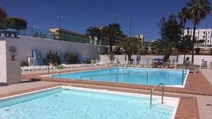Två rektangulära pooler med kakelbeklädda kanter, omgivna av vita solstolar och parasoller, med palmer och byggnader som bakgrund.