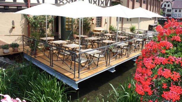 Un café al aire libre con mesas y sillas, rodeado de plantas en macetas y una terraza de madera sobre el agua.