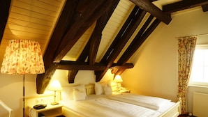 Un dormitorio con techo inclinado, dos mesitas de noche con lámparas, una cama con ropa blanca y un ventanal con cortinas.
