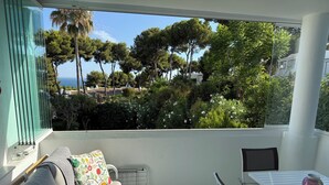 Ein Balkon mit einer Couch, einem Tisch und Stühlen, der auf einen Garten mit Bäumen und Meerblick führt.