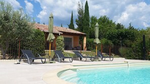 Zona de piscina con sillones, sombrillas y un edificio con techo de tejas.