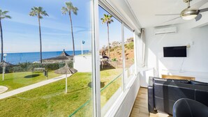 Une chambre dotée d’une grande fenêtre donnant sur une plage, d’un ventilateur de plafond et d’un téléviseur à écran plat fixé au mur.