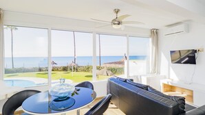 Un salon moderne doté d’une grande fenêtre offrant une vue sur la mer et une piscine. On y trouve un ventilateur de plafond, un téléviseur à écran plat et un coin repas avec une table et des chaises.