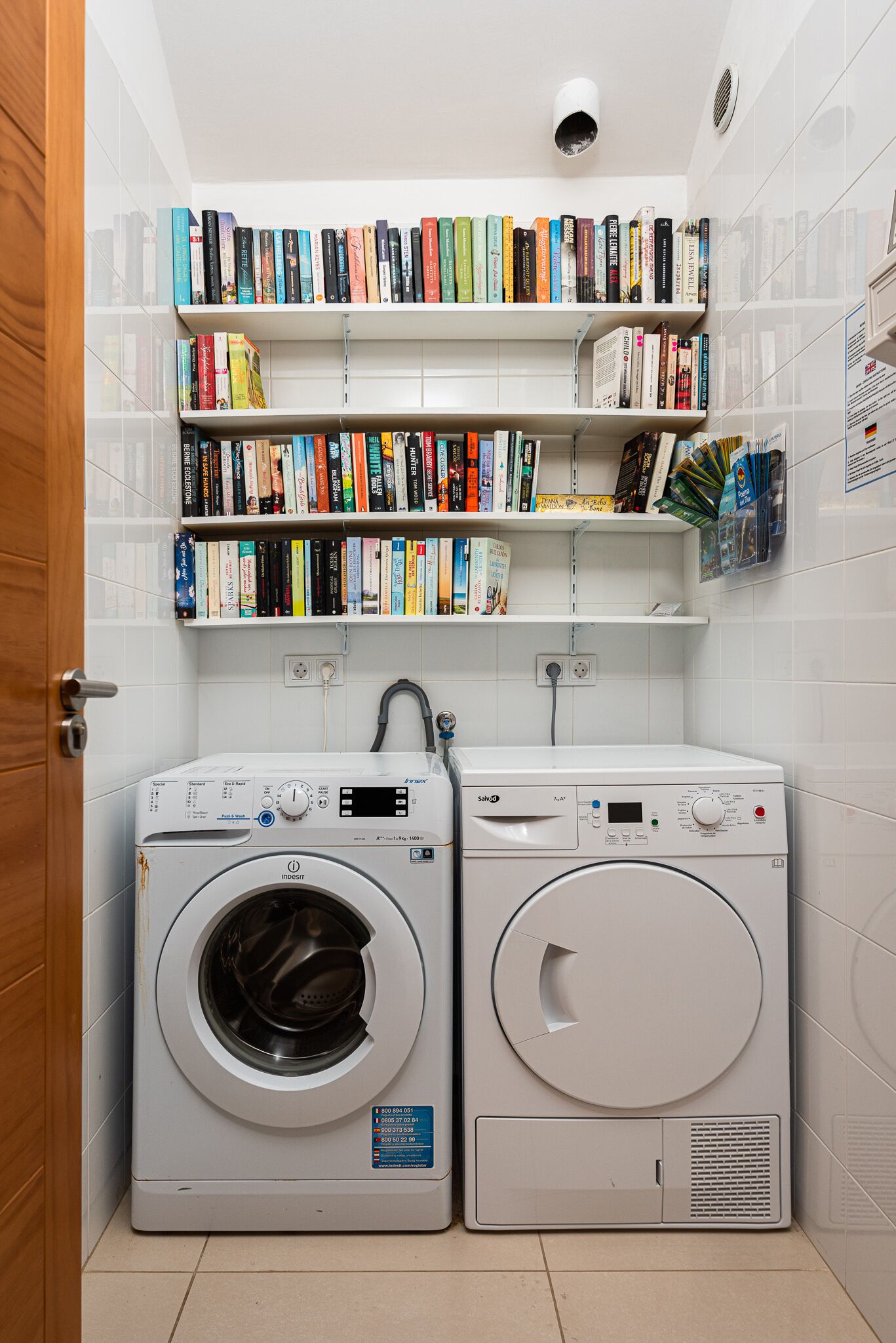Un cuarto de lavandería con dos lavadoras, una estantería con varios libros y una pared de azulejos.