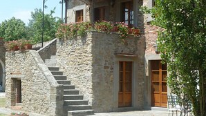 Ein Steinhaus mit einer Treppe zu einer Holztür, einem überdachten Außenbereich mit Metallrahmen und einem Garten mit Topfpflanzen.