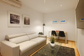 Ein modernes Wohnzimmer mit einem beigen Sofa, einem Glastisch und einem Schreibtisch mit zwei Stühlen. An den Wänden hängen gerahmte Bilder und es steht eine Stehlampe im Raum.