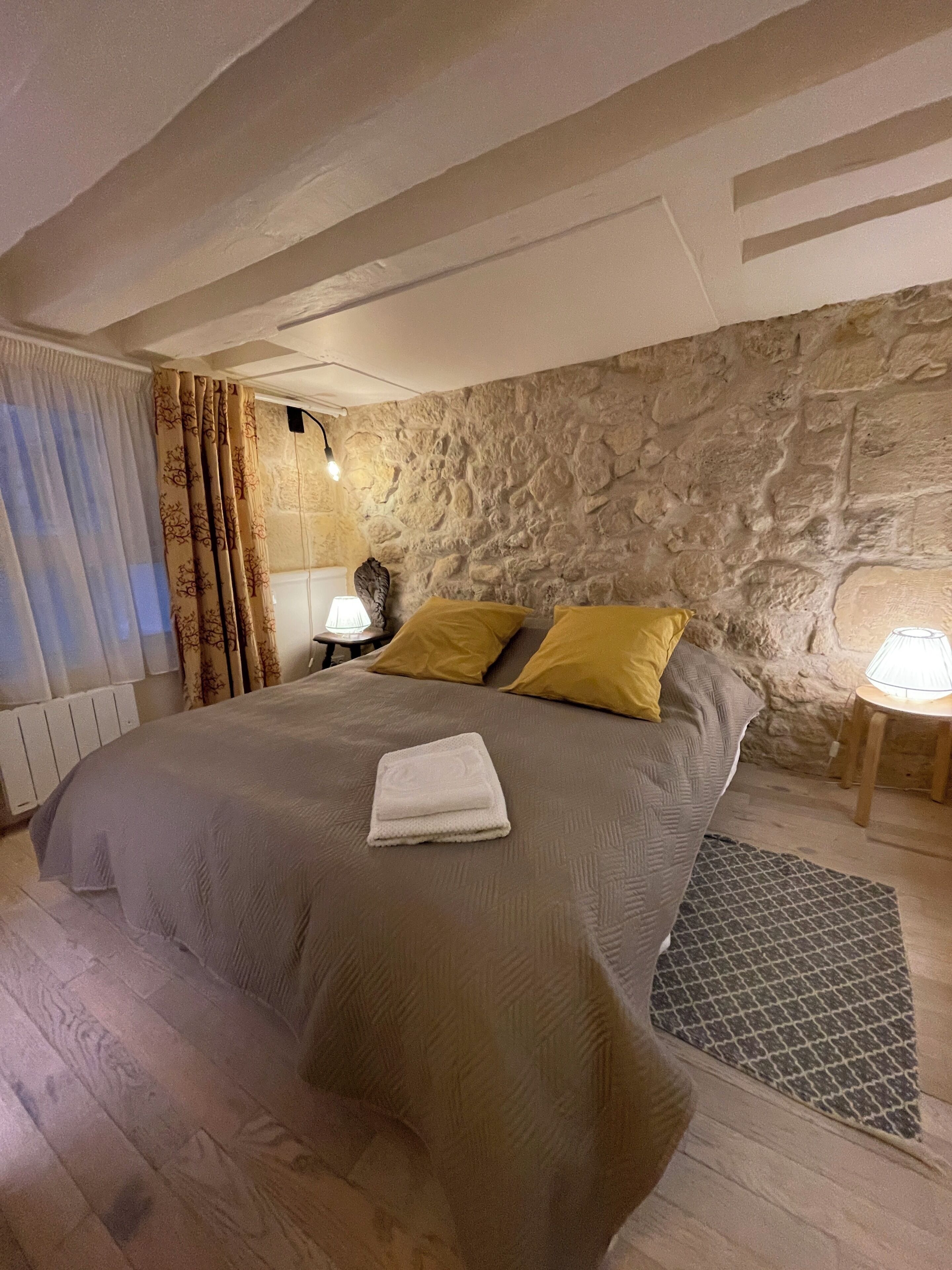 A bedroom with a stone wall, a bed with a grey bedspread and two yellow pillows, a bedside lamp, and a folded towel on the bed.