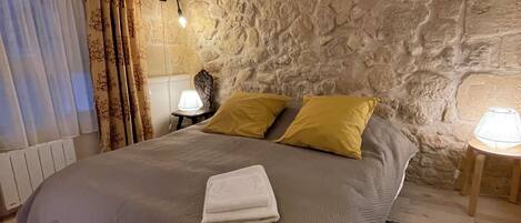 A bedroom with a stone wall, a bed with a grey bedspread and two yellow pillows, a bedside lamp, and a folded towel on the bed.
