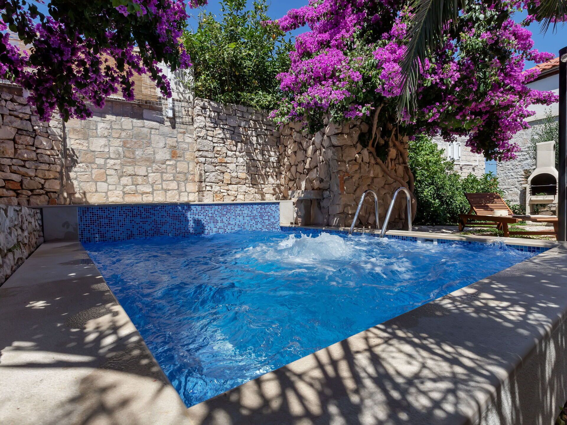 A swimming pool with stone walls and a tiled edge, surrounded by lush greenery and purple flowers.