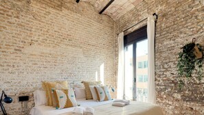 A bedroom with a brick wall, a bed with white bedding and decorative pillows, a bedside table with a lamp, and a window with curtains.