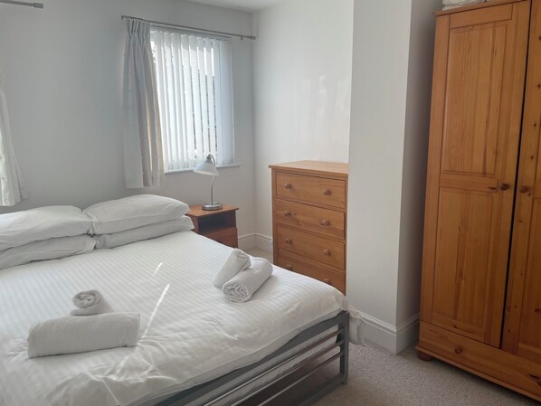 Une chambre à coucher avec un lit, une commode en bois, une table de chevet avec une lampe et une fenêtre avec des stores.