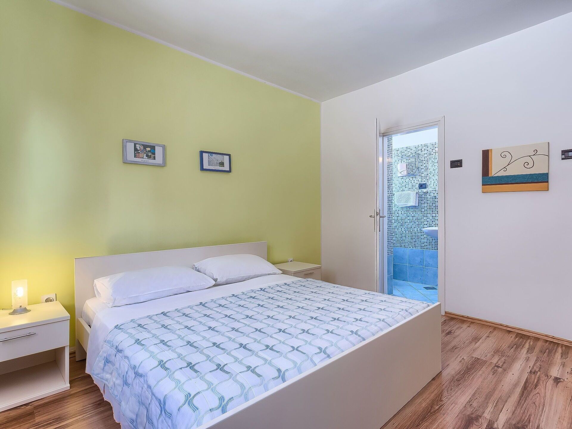 Ein Schlafzimmer mit einem Bett, einem Nachttisch, einem Fenster mit Blick auf ein Gebäude und zwei gerahmten Bildern an der Wand.