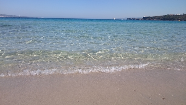 Une plage avec une eau claire et peu profonde, un rivage sablonneux et, au loin, une terre ferme parsemée de bâtiments.