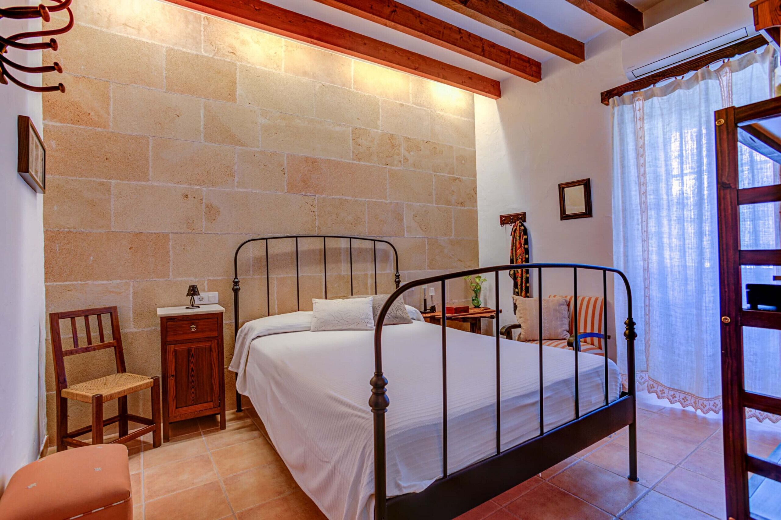 A bedroom with a metal bed frame, a wooden nightstand, a wicker chair, a small cabinet, and a window with curtains.
