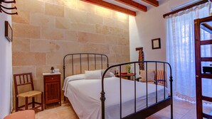 Une chambre à coucher avec un lit en métal, une table de chevet en bois, une chaise en osier, une petite armoire et une fenêtre avec des rideaux.