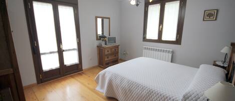 A bedroom with a large bed, wooden floor, a dresser, a mirror, and a window with curtains.