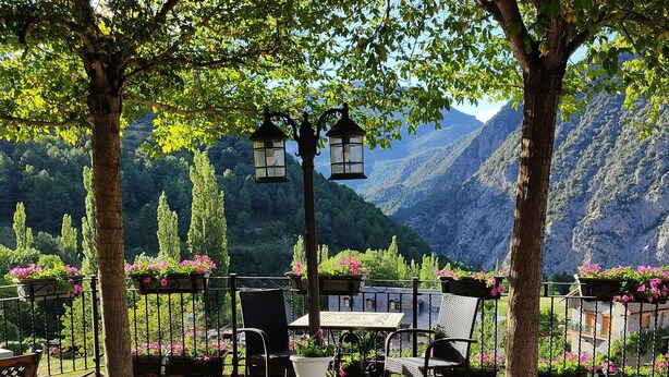 Ein Außenbereich mit Tischen und Stühlen, umgeben von Bäumen und Blumenkästen, mit Blick auf eine Berglandschaft.