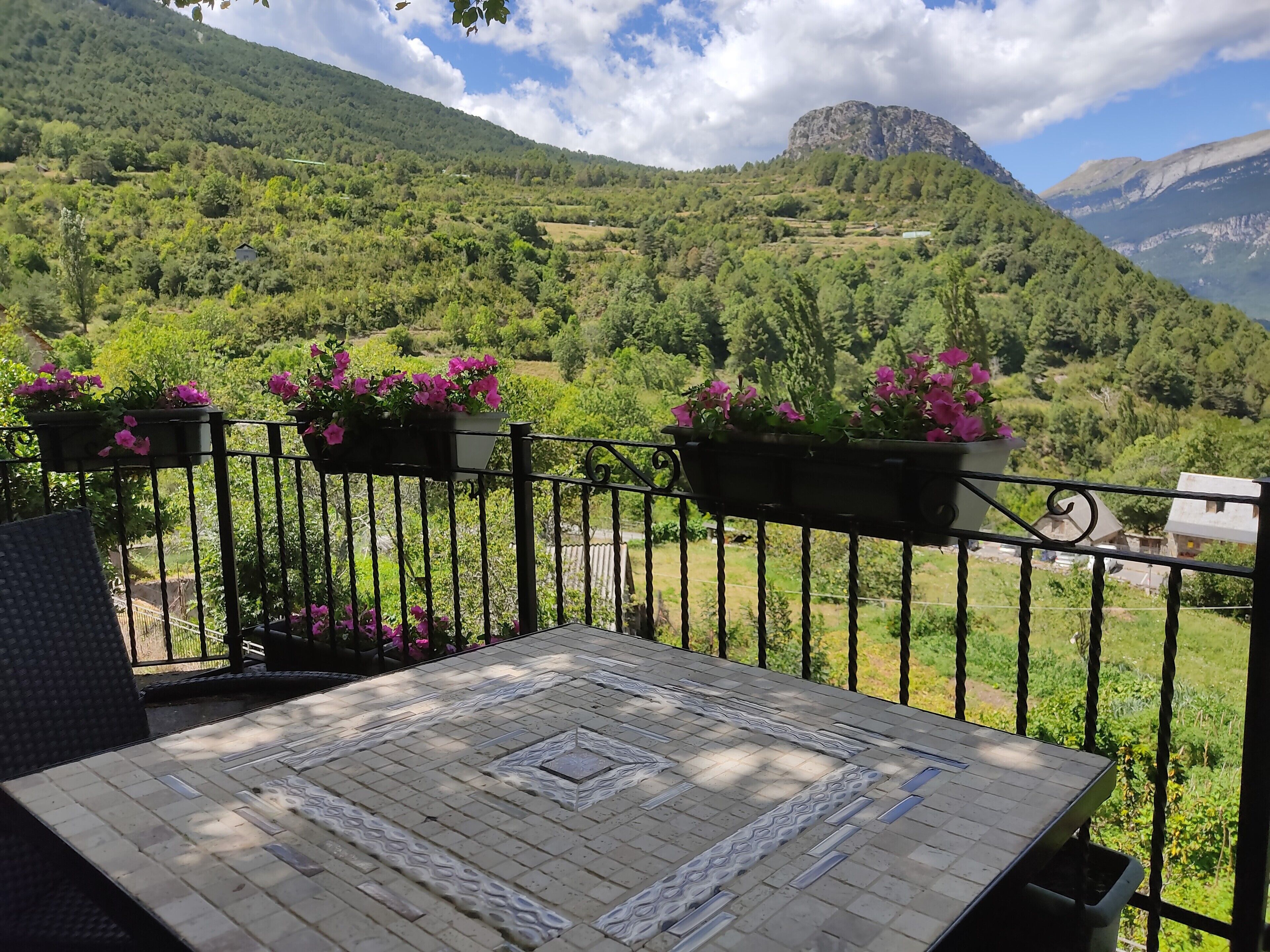 A balcony with a view of a mountainous landscape, a table with a patterned tablecloth, and flower planters.