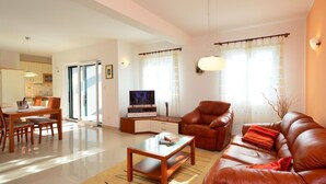 Una sala de estar moderna con un sofá de cuero, una mesa de centro de madera y un televisor sobre un mueble de madera. Se observa un comedor con una mesa y sillas, y una cocina al fondo.