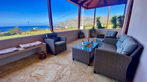 A covered outdoor seating area with wicker furniture, a stone floor, and a view of the ocean and hills.