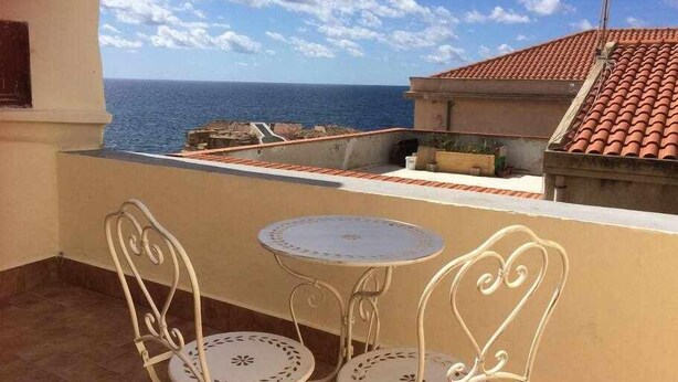 Ein Balkon mit zwei weißen Metallstühlen und einem kleinen runden Tisch, mit Blick auf eine Küstenlandschaft, strahlend blauer Himmel und das Meer im Hintergrund.