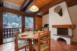 Een houten interieur met een eettafel gedekt voor twee personen, een open haard en uitzicht op besneeuwde bomen door grote ramen.