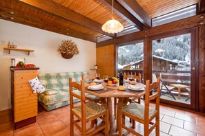Un coin repas chaleureux avec une table en bois dressée pour deux personnes, un banc rembourré et une vue sur un paysage enneigé par de grandes portes vitrées.