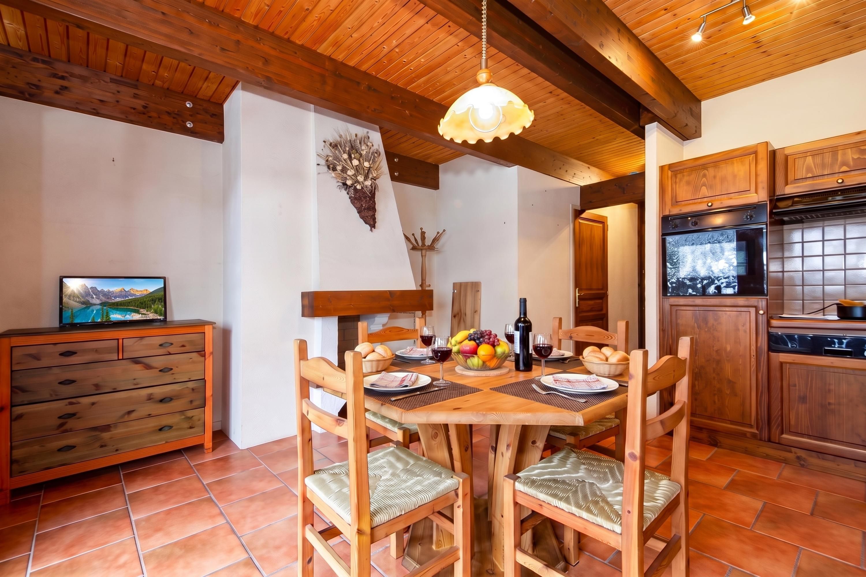 Une table de salle à manger en bois, avec des chaises, un panier de fruits, des verres à vin et une bouteille de vin.