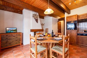 Een houten eettafel met stoelen, een schaal met fruit, wijnglazen en een fles wijn.