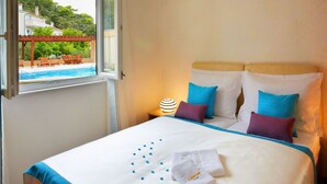 A hotel room with a bed, pillows, a nightstand, a lamp, and a window with a view of a pool and greenery.