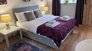A neatly made bed with a purple blanket, two pillows, and two folded towels. There is a bedside table with a lamp, a rug, and a window with curtains. - Stables Retreat - Direct Access to The Forest (Burley)