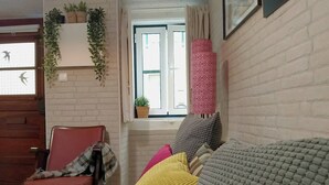 A living room with a red chair, a sofa with patterned cushions, a window with curtains, and a brick wall. - Vintage Apartment in Anjos, Central Lisbon (Lisboa)
