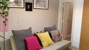 A living room with a white brick wall, a sofa with multiple cushions, a small plant, and framed artwork. - Vintage Apartment in Anjos, Central Lisbon (Lisboa)