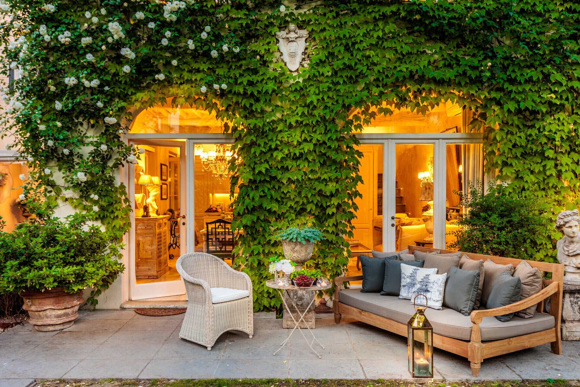 An outdoor seating area with a wicker chair and a sofa, surrounded by lush greenery and a stone statue.