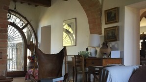 Une pièce chaleureuse avec une table en bois, un fauteuil en cuir, un canapé et un coin repas comprenant une table et des chaises. Il y a une fenêtre donnant sur des arbres et un mur en briques.
