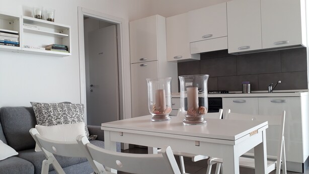 Une cuisine moderne avec des meubles blancs, une table à manger avec des chaises et un canapé gris agrémenté de coussins à motifs.
