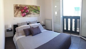 Une chambre à coucher comprenant un lit, des oreillers, une table de chevet, une lampe, un radiateur et une porte-fenêtre coulissante donnant sur un balcon.