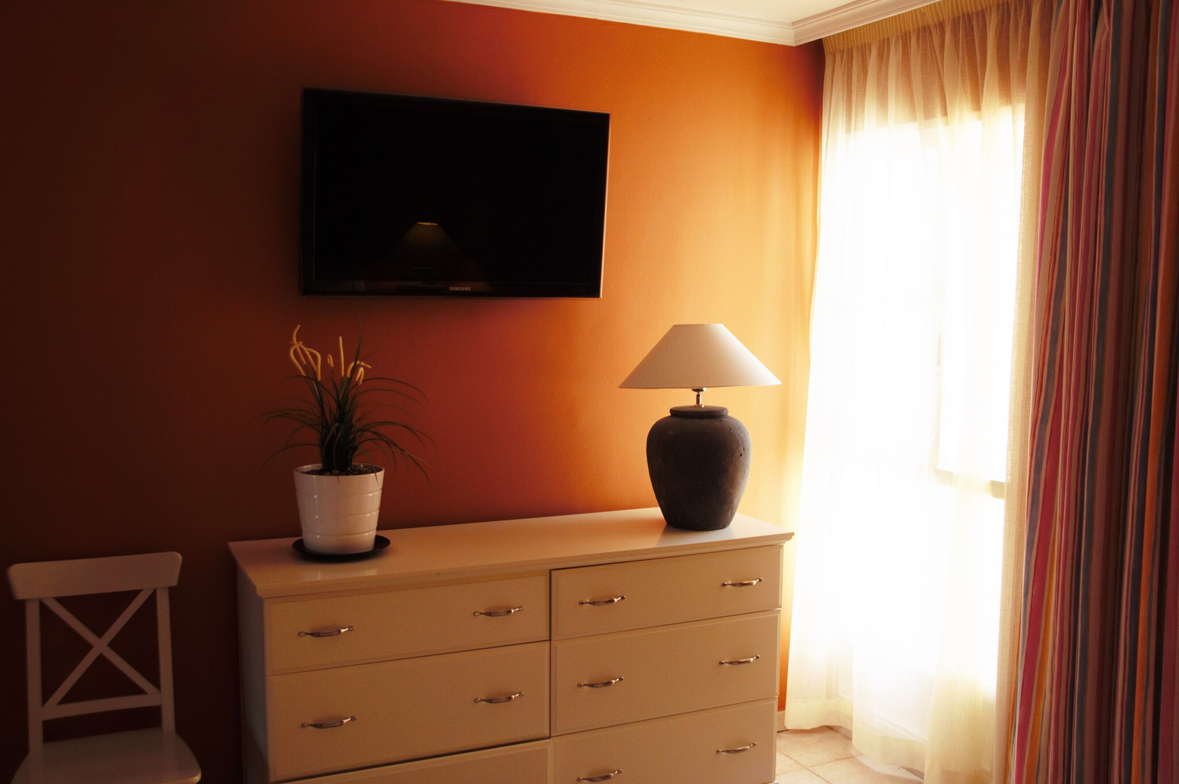 A room with a TV mounted on the wall, a lamp on a dresser, and a potted plant.