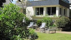 Une maison à deux étages dotée d’un balcon, entourée d’une végétation luxuriante et agrémentée d’une petite terrasse avec des meubles d’extérieur.
