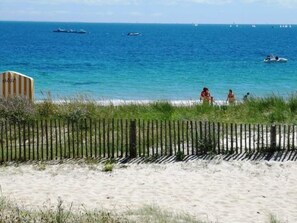 A beach with a wooden fence, sandy shore, and a clear blue sea with boats.