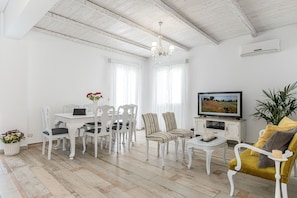 Un salon spacieux avec un ensemble de salle à manger blanc, un fauteuil jaune, un téléviseur et une grande fenêtre avec des rideaux.